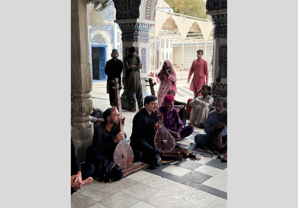 A group of people sitting on the floor of a beautifully tiled shrine, with two men playing traditional stringed instruments, surrounded by others listening attentively.